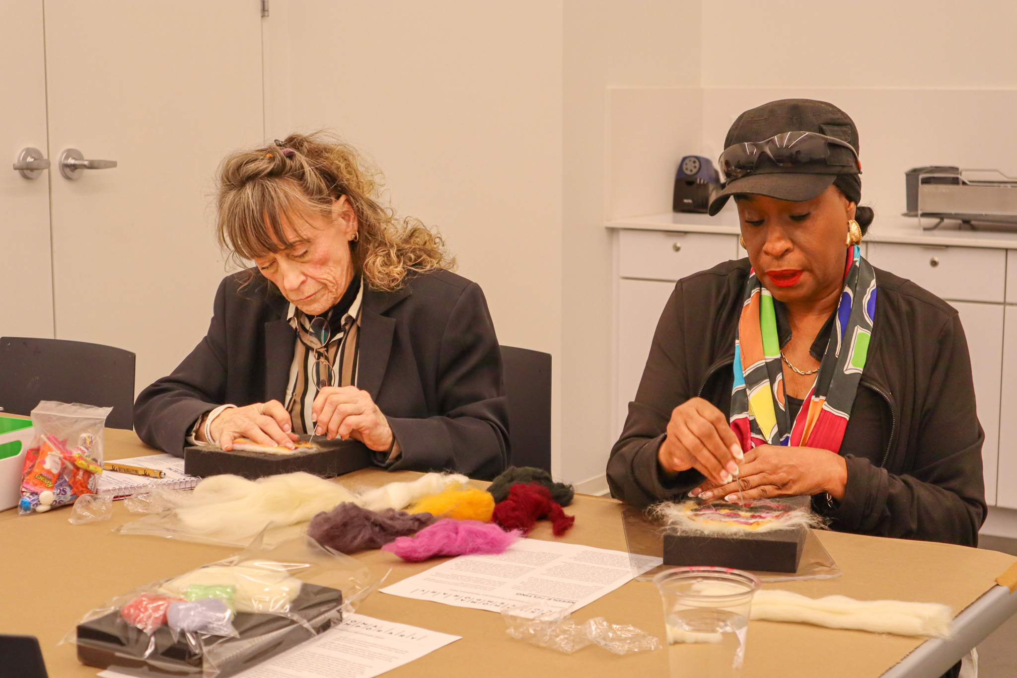 An older white and Black woman sit next to each other at a table, surrounded by colorful wool. They are both focused working on a felting project.