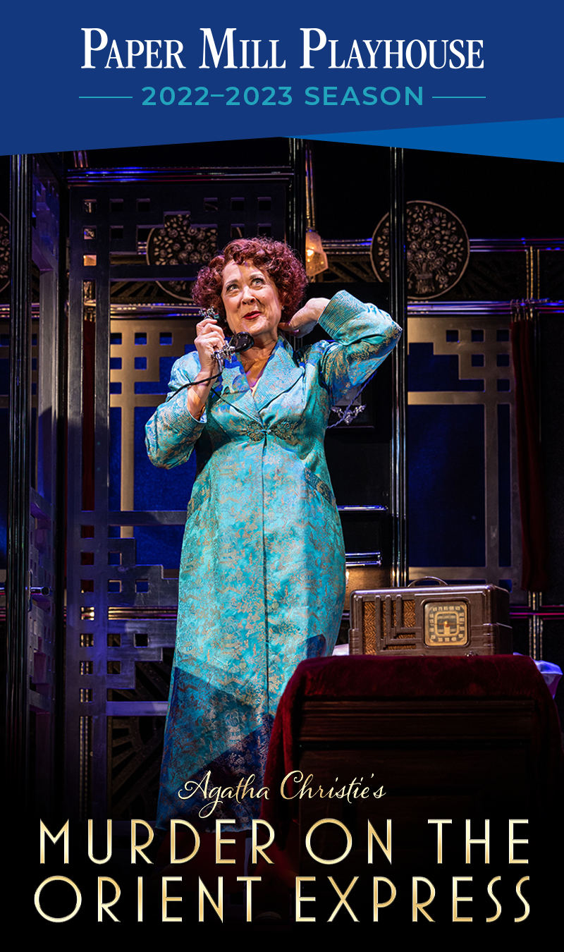 A blue border with white and light blue text reads, "Paper Mill Playhouse, 2022-2023 Season" above a photo of an actress in Murder on the Orient Express standing in the middle of the stage set of a lavish train compartment, dressed in a blue dressing gown, speaking on a rotary telephone, and posing pleasantly with one hand fluffing her hair. At the bottom of the photo, a title reads, "Agatha Christie's Murder on the Orient Express."