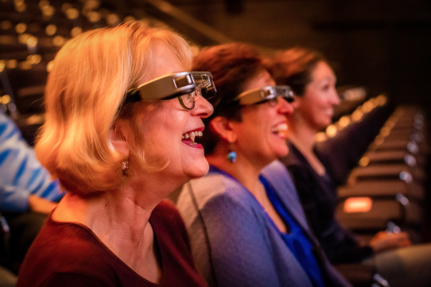 Three laughing women sit in the Steinbright Theatre while wearing Smart Caption Glasses