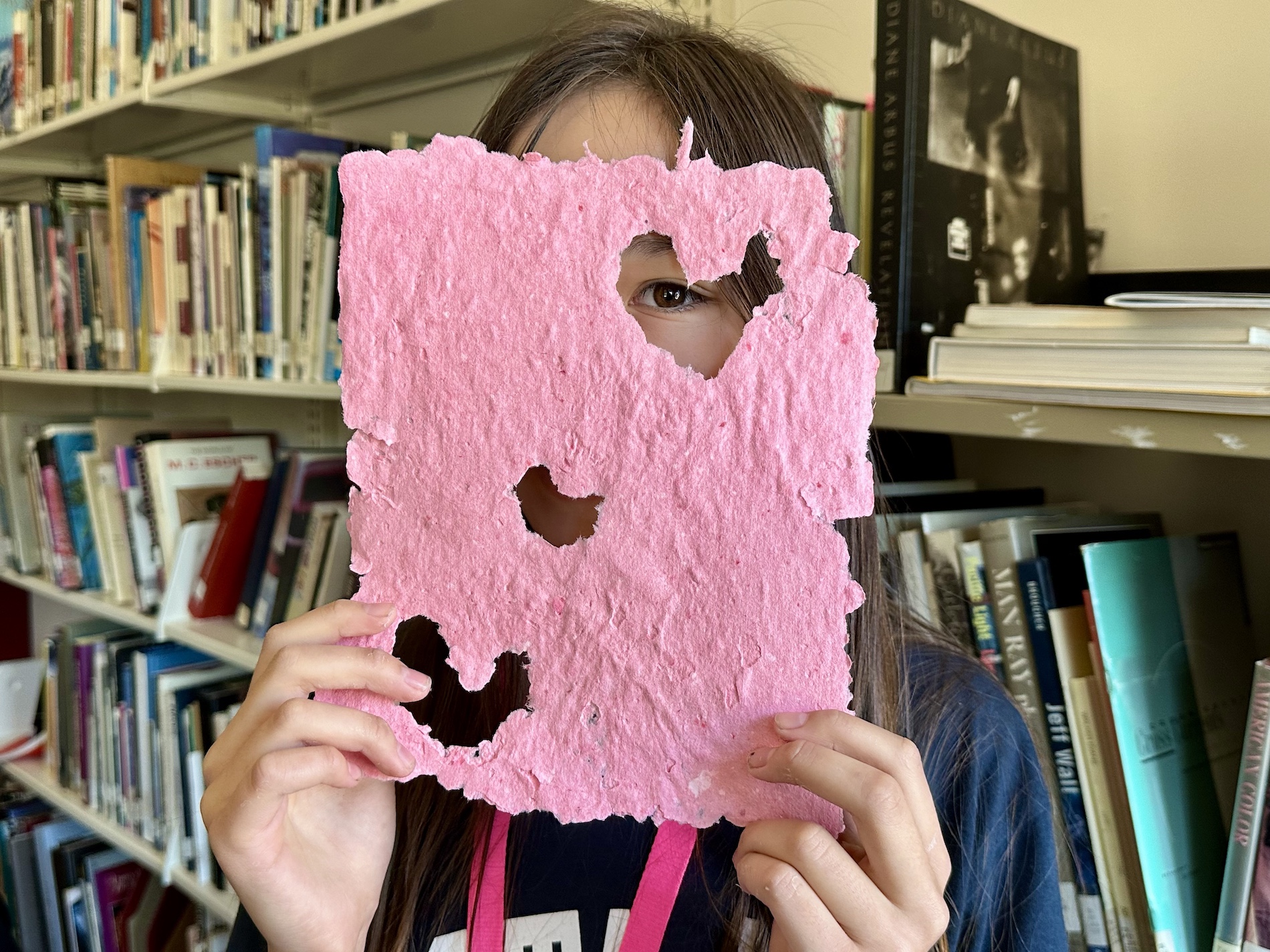 A teen holds a sheet of pink handmade paper, peering through a heart-shaped cutout with one eye.