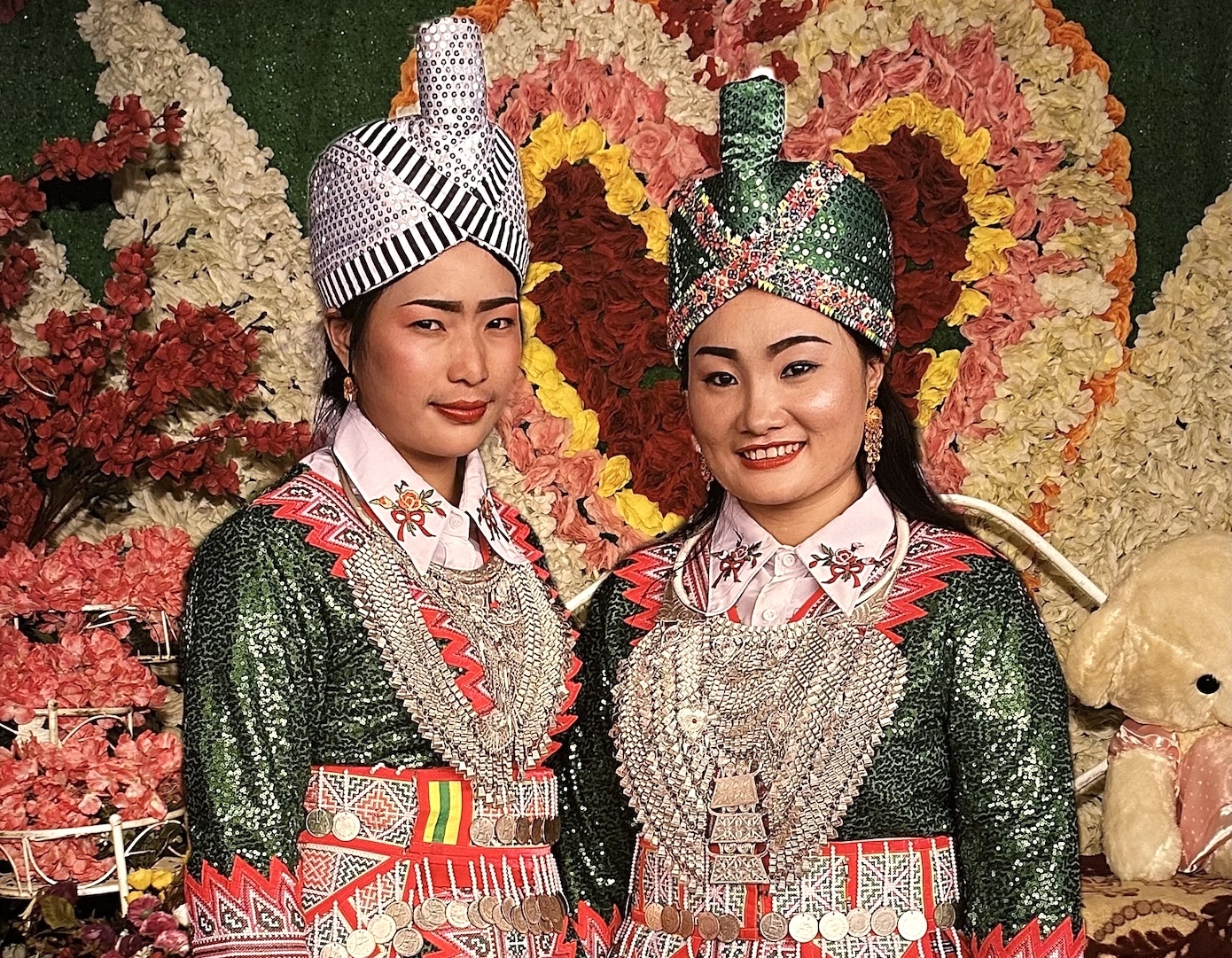 Two women pose in front of a floral heart arrangement wearing traditional Hmong outfits with embroidered jackets, elaborate hats, and silver jewelry.