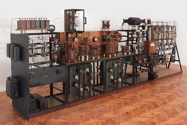 A large black and brown contraption in a white room with brown flooring. This is a transmitter and has many dials and wires.