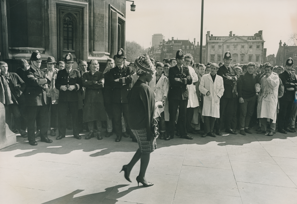 Photograph of a Black woman walking past a group of white men, police officers lining the road. This photograph was taken outside the House of Commons. The men are there to demonstrate their support for the racist and fascist MP, Enoch Powell.