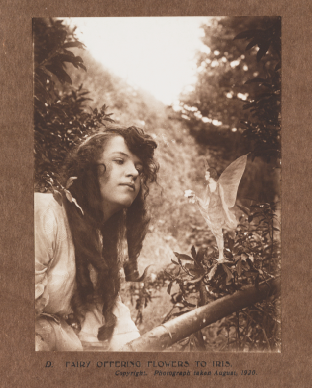 A young girl seems to interact with a small fairy against a woodland background. The image is captioned 'Fairy offering flowers to Iris' and is one of the set of a few staged photos from the Cottingley Fairies scandal in the early twentieth century.