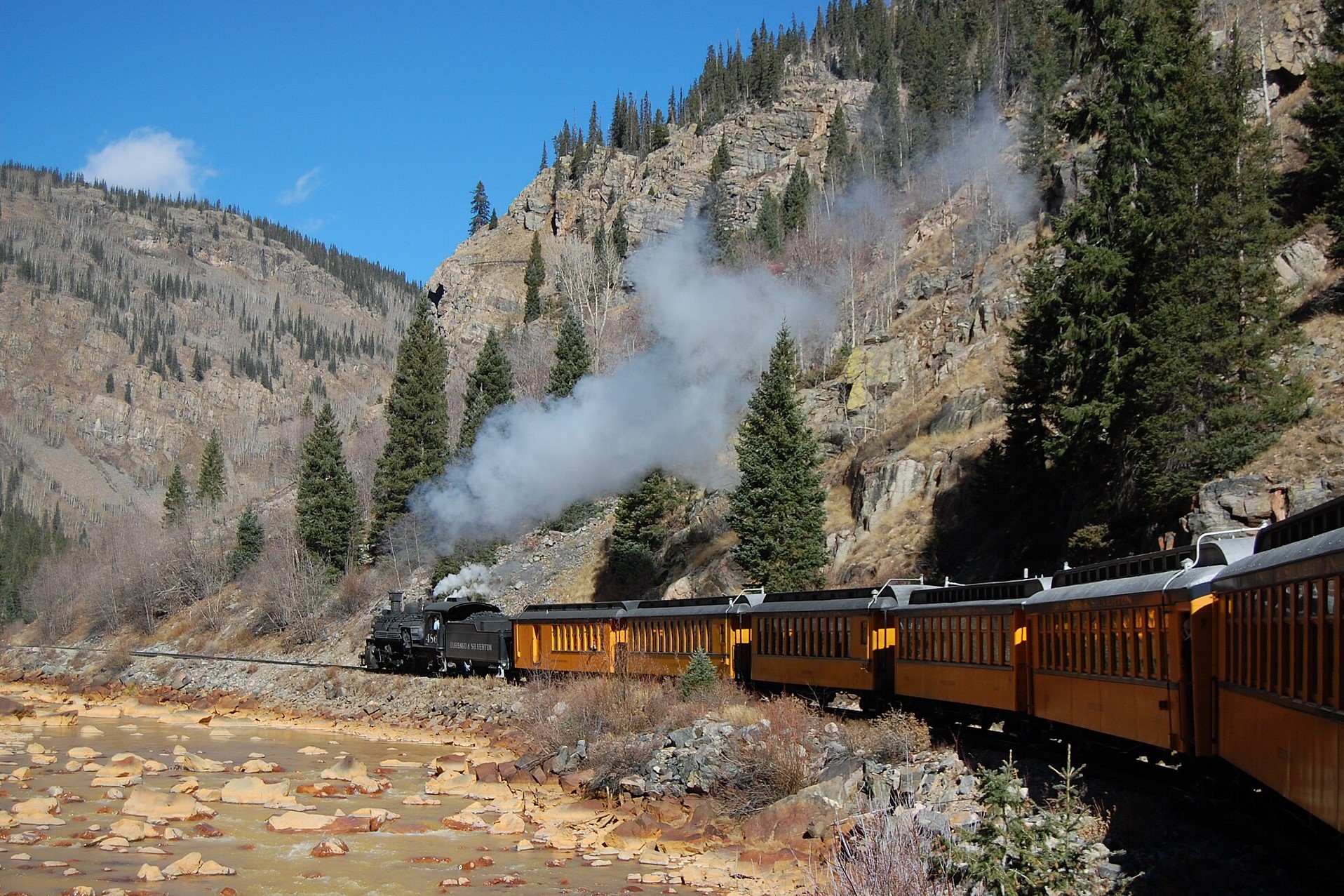 Image of a steam train moving away from the viewer along a rocky, tree-studded hillside. The train's cars are yellow and a shallow, rocky body of water sits to the left.