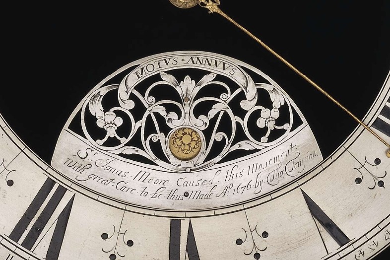 Very close up image of a black and white clock face, with roman numerals across the bottom and a long golden hand. A golden flower sits above the inscription.
