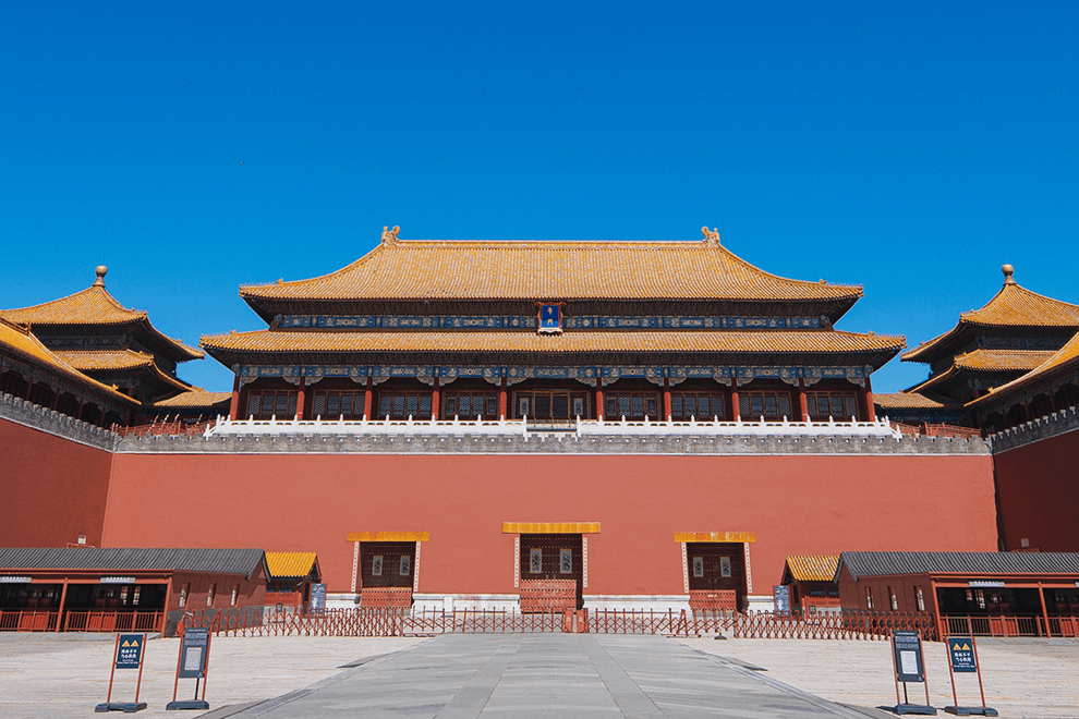 Image of the Palace Museum in Beijing, featuring a red wall and two curved roofs, against a bright blue sky.