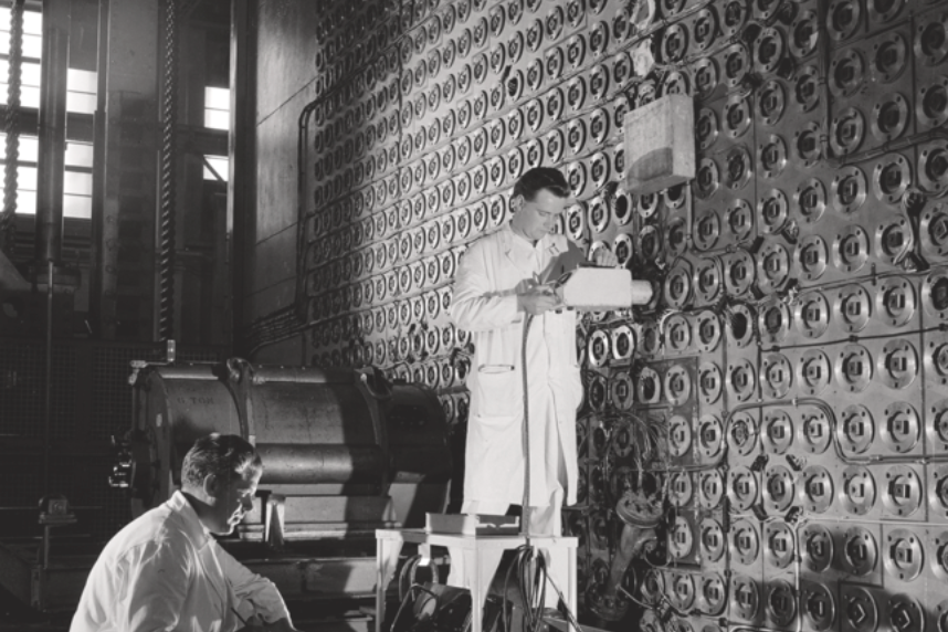 Black and white image of two scientists, young men in white coats, examining a wall full of round windows.