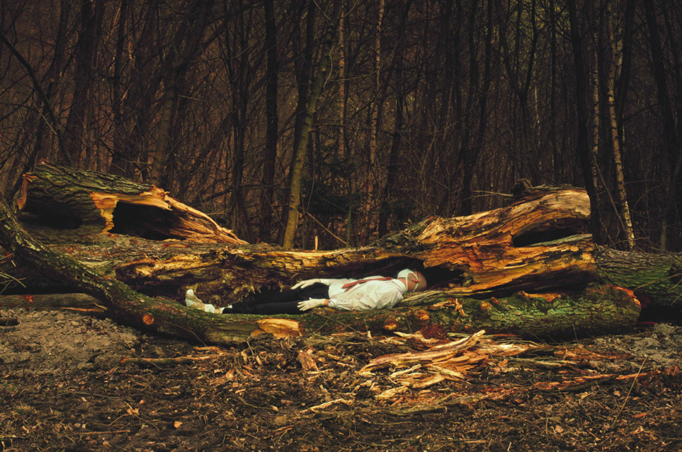 Photograph of ill person resting in splintered fallen tree against dark forest background. Václav Jirásek, Infection (2022, London: Eastern Front).