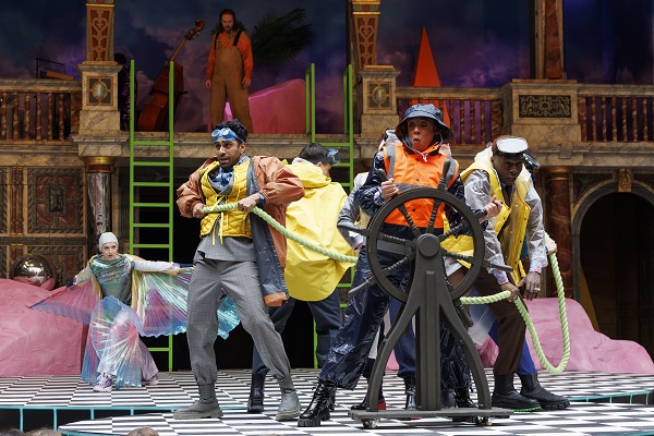 [IMAGE] A group of actors wearing lifejackets are crowded around a ship's wheel on a chequerboard stage.