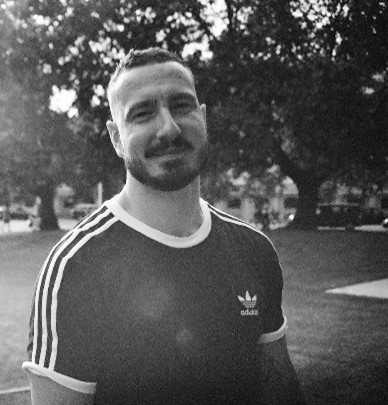 [Image] A black and white photo of HE team member Craig Ritchie in a park. He is wearing a black t-shirt and smiling.