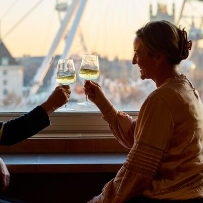 Two people seated in the Members' Lounge by a window with a view of the London Eye clinking wine glasses together whilst smiling.