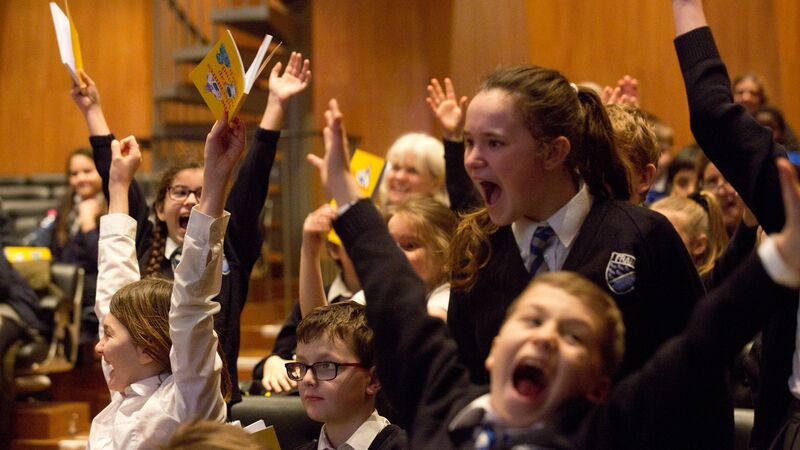 Excited children putting their hands up and smiling in an auditorium. 