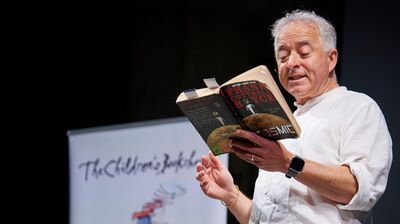 Frank Cottrell-Boyce in a white shirt reading from a book onstage.