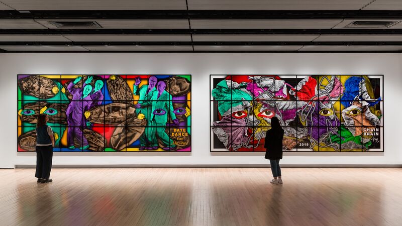 Two people looks at two large and vibrant artworks hung up in a large space with white walls and wooden floors.