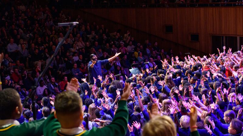 School children in different uniforms on stage with a choir leader facing them with arms raised.