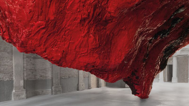 Large red texture sculpture hanging down from the ceiling to the floor in a space with stone floors and pillars
