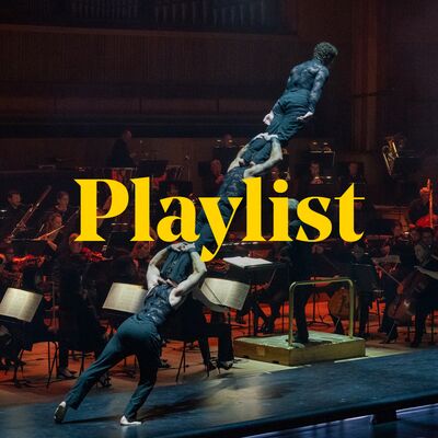 A quartet of acrobats, clad in back, lean forwards while standing on top of each others' shoulders. Behind them, on the same stage, an orchestra performs. The word 'Playlist' is imposed over the top of the image.