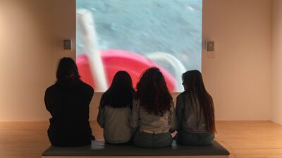 A group of students sat on a bench inside the Hayward Gallery facing a wall where a video is playing.