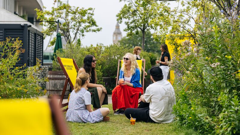 Friends relax on a grassy rooftop terrace with yellow deckchairs, featuring Big Ben and the London Eye in the background.