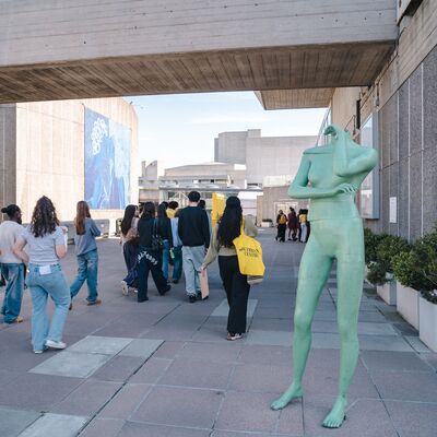 A tall, mint-green, headless female statue stands on a paved plaza as a group of people walk beneath a concrete walkway.