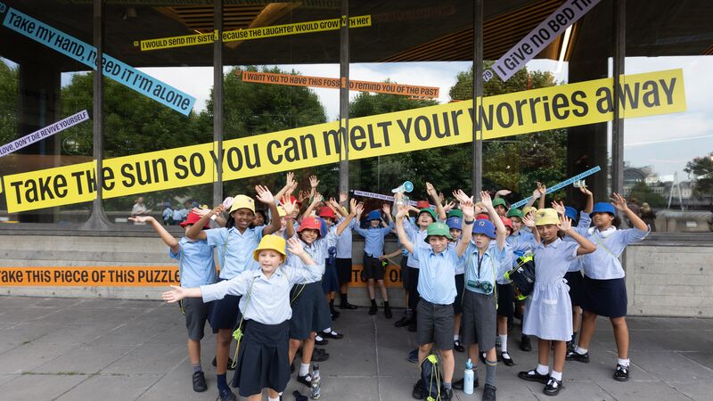 A group of schoolchildren with their arms in the air with the Gift to the Future poetry lines behind them