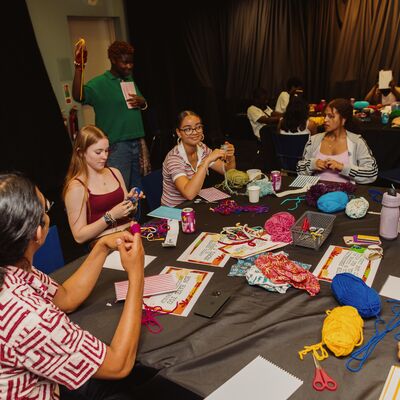 A group of young people and an instructor sit around a large black table covered in colorful skeins of yarn, paper handouts, and crafting supplies during a workshop.
