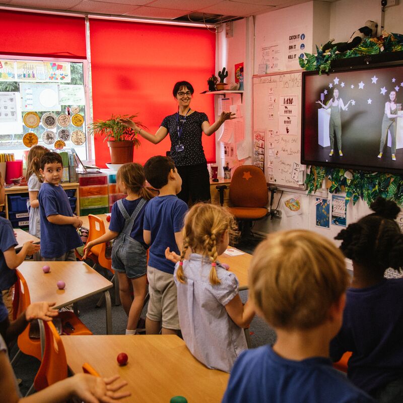Small school children stand at their desks as their teacher leads a music class