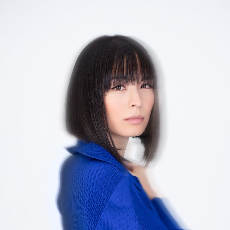 Alice Sara Ott is standing in front of a white background. She is turned towards the camera in a 3/4 pose, with a hand up to her chest. She looks into the camera. She is wearing an electric blue blouse.