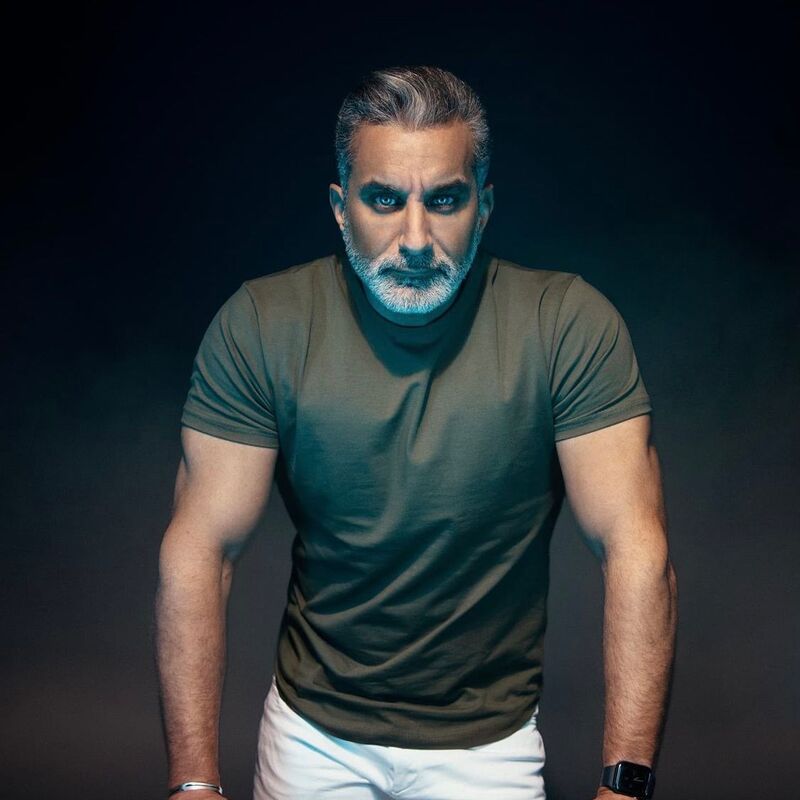 Bassem leans forward placing his hands on a low black table and looks imperiously straight to camera. He wears white jeans and a green top. He has grey hair and a neat white beard.