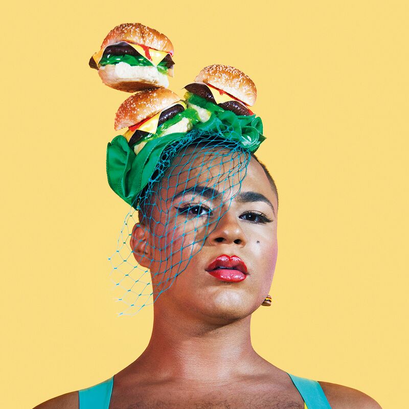 Performance artist Travis Alabanza wearing a fascinator headpiece, a layer of 3 cheeseburgers on top