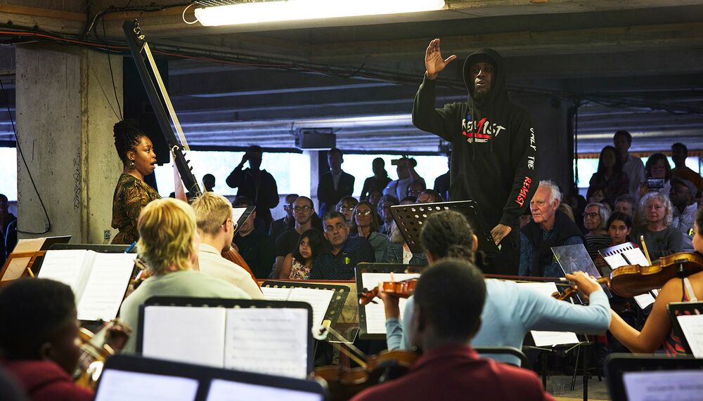 Moustapha Doumbia conducts the Multi-Story Orchestra in a multi-story car-park; behind Doumbia an audience watches on.