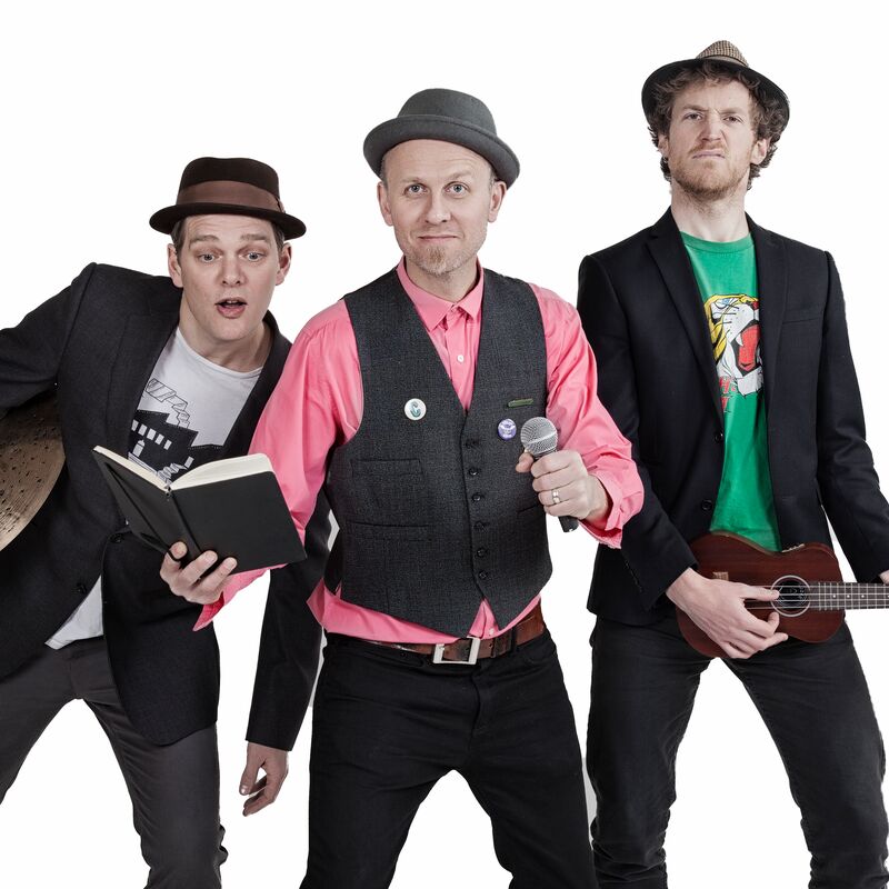 3 men stand in a row in various poses. The man on the left of the image holds a cymbal under his arm and leans to the right to see the shoulder of the man in the middle. He wears a hat, grey trousers, and a black blazer over a white t shirt. The man in the middle of the image wears a grey hat, a pink shirt, a grey waistcoat and black jeans with a brown belt. In his left hand he holds a microphone and in his right hand he holds a notebook open. The final man on the right of the image stands slightly taller than the other two. He also wears a hat and blazer but with a green t shirt underneath. He poses as though he is strumming his ukulele.