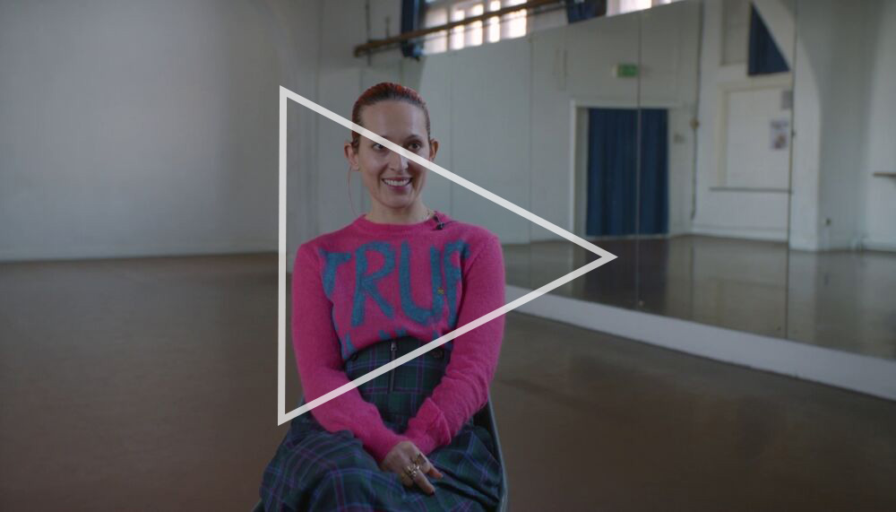 Holly Blakey sits in an empty dance rehearsal studio. She has slicked back red hair and wears a pink sweater with the word 'TRUE' in large letters on the front. Behind her we can see a large mirrored wall reflecting the rest of the studio space