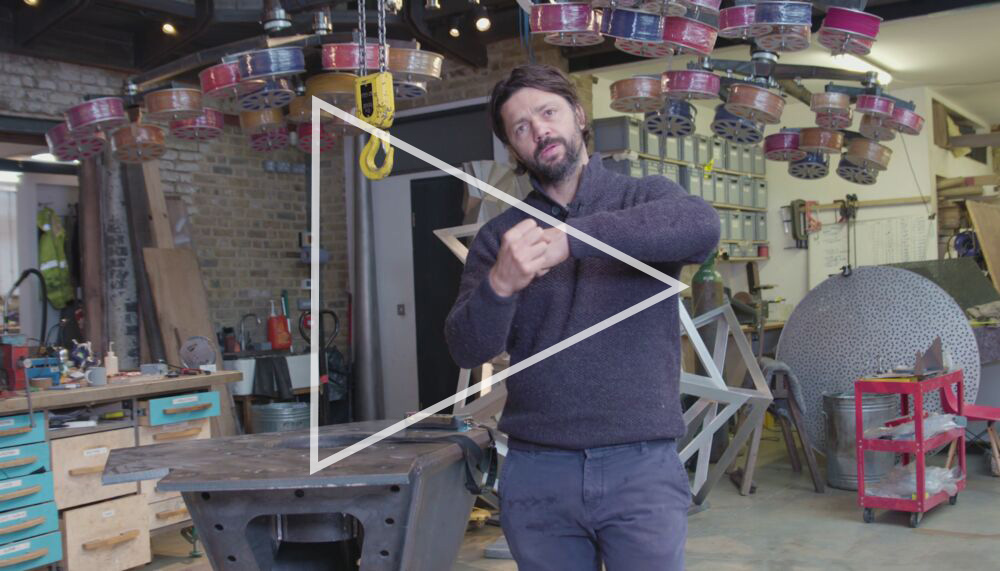 Artist Conrad Shawcross stands in his studio and workshop with his hands clasped in a demonstration; behind him are various tools and machines, a set of drawers, and many fabricated art pieces