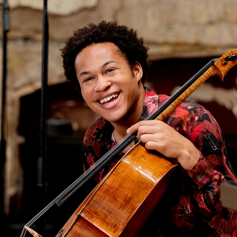 Cellist Sheku Kanneh-Mason wearing a black and burgundy patterned shirt, smiling, holding a cello