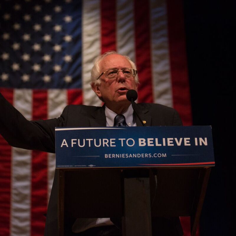 Senator Sanders stands infront of an American flag gesturing upwards with his right arm. He has white hair and glasses and the lectern he stands behind bears the sign 'A Future To Believe In'