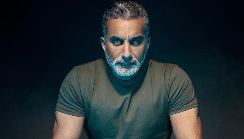 Bassem leans forward placing his hands on a low black table and looks imperiously straight to camera. He wears white jeans and a green top. He has grey hair and a neat white beard.