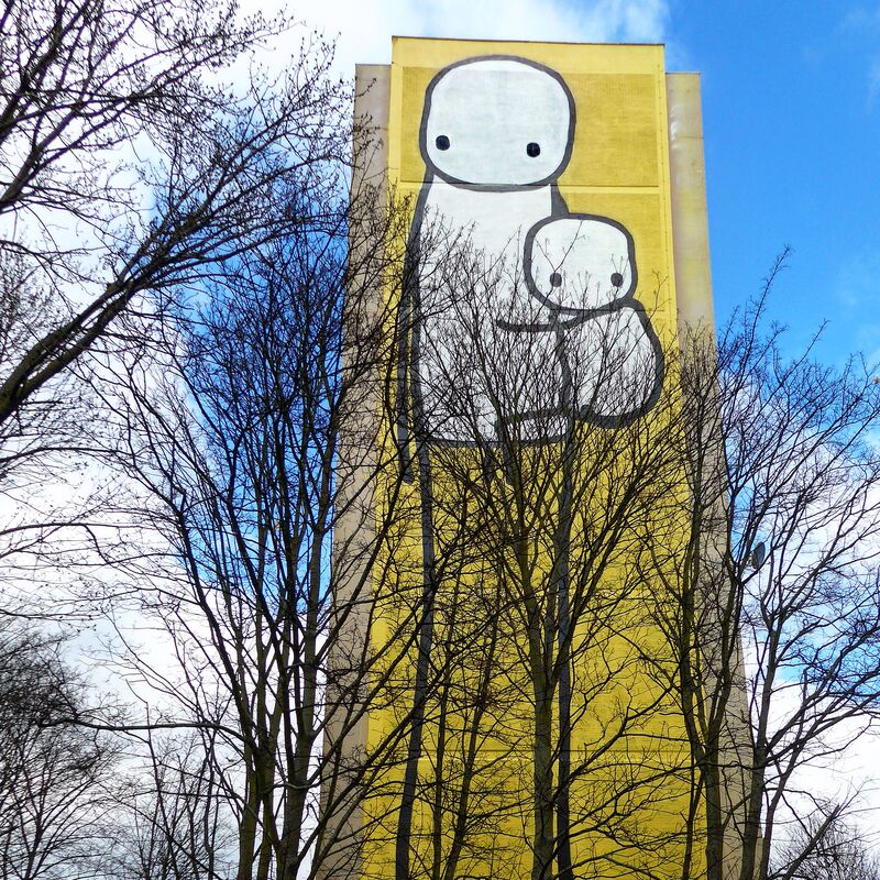 A photo of a very tall yellow building with STIK artwork on of a large stik figure holding a baby stik figure