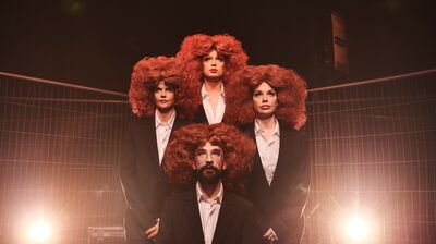 Four performers in black suits and white shirts all wear large red curly wigs