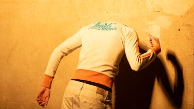 Someone wearing a white jacket and jeans leans their head against a cream wall