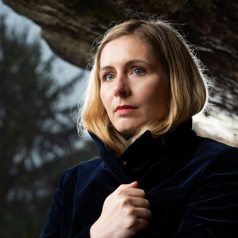 A white woman with blonde hair holds her blue wool coat closed at her chest. She looks off camera against a backdrop of trees,