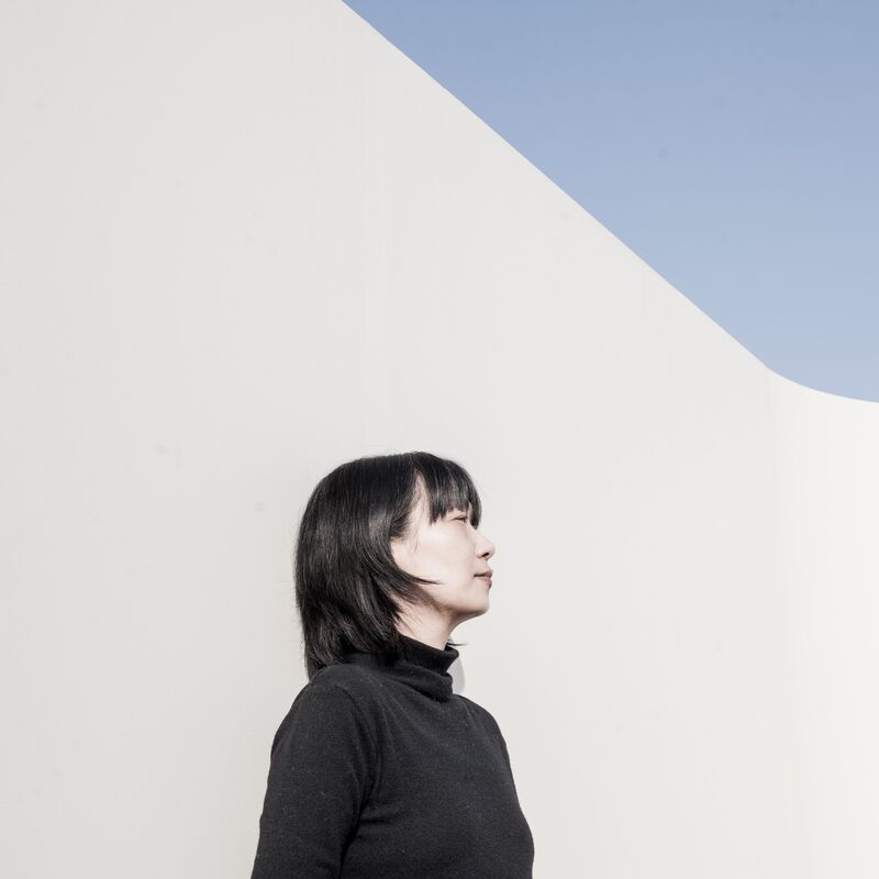 Han wears a black turtleneck and is pictured in side-profile. She's got cropped black hair and a fringe and is pictured outdoors standing against a white wall. Blue sky is visible in the top right corner of the image.