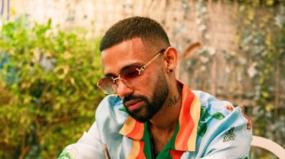 Artist Jaz Dhami in a colourful shirt and sunglasses, sat at a table outside.