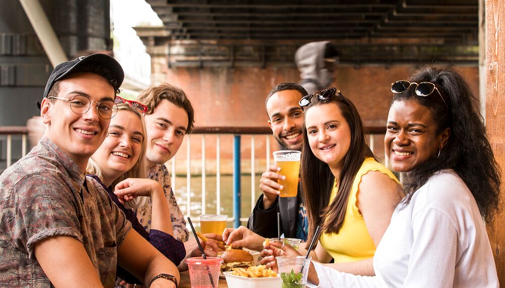 A group of six young people sit at an oudoor table enjoying food and drink