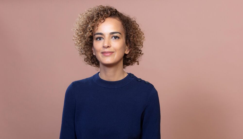 Leila has cropped, curly honey coloured hair and smiles to camera. She wears a blue jumper and is standing against a pink/brown background.