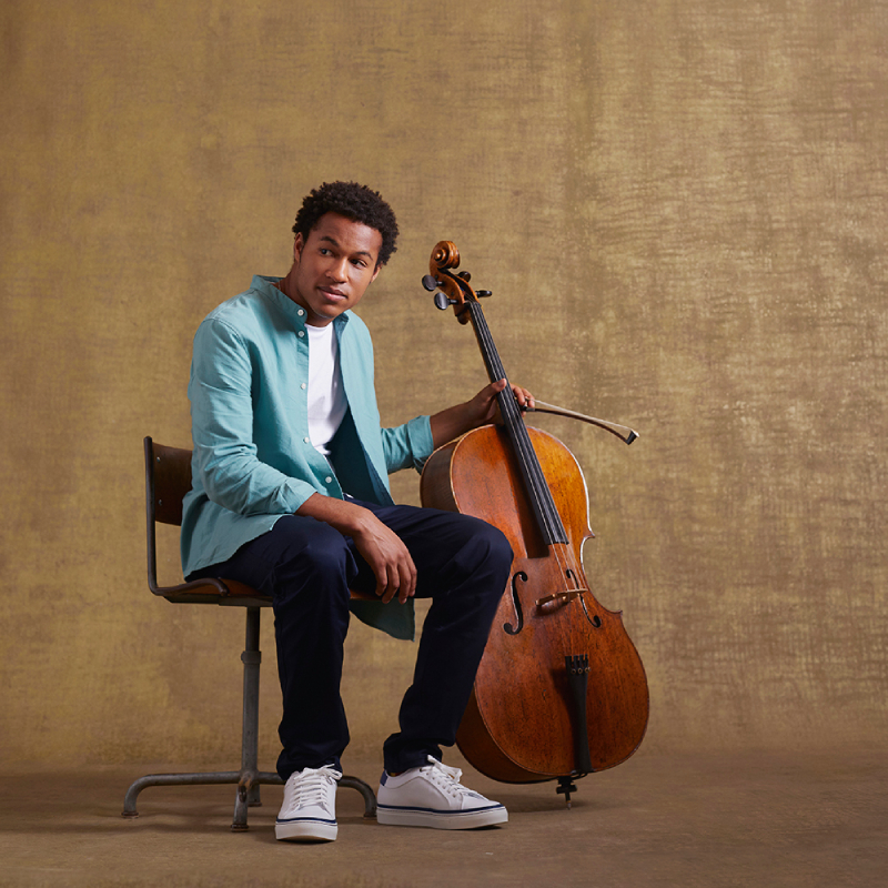 Cellist Sheku Kanneh-Mason in a denim shirt casually seated with his cello