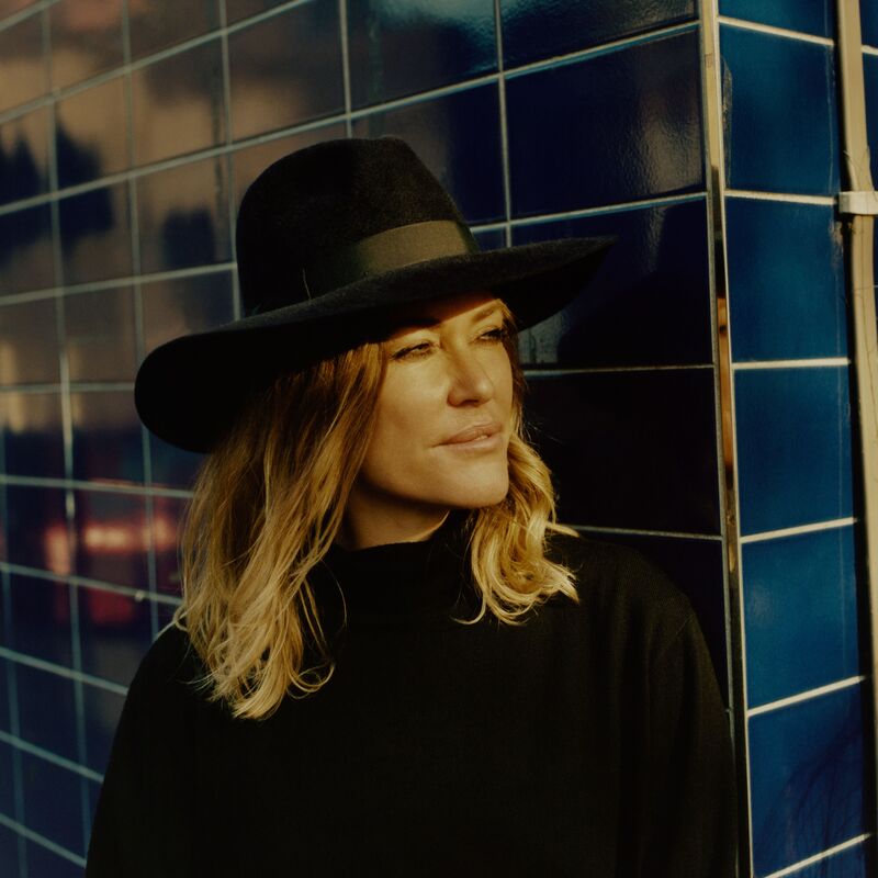 Cerys Matthews stands against a blue tiled wall wearing a black wide-brimmed hat