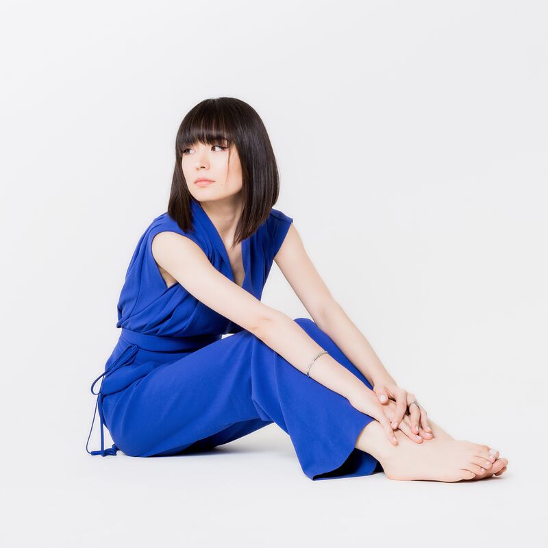 Alice Sara Ott is wearing a bright blue jumpsuit. She sits on a neutral white background, her hands resting on her ankles and bare feet.