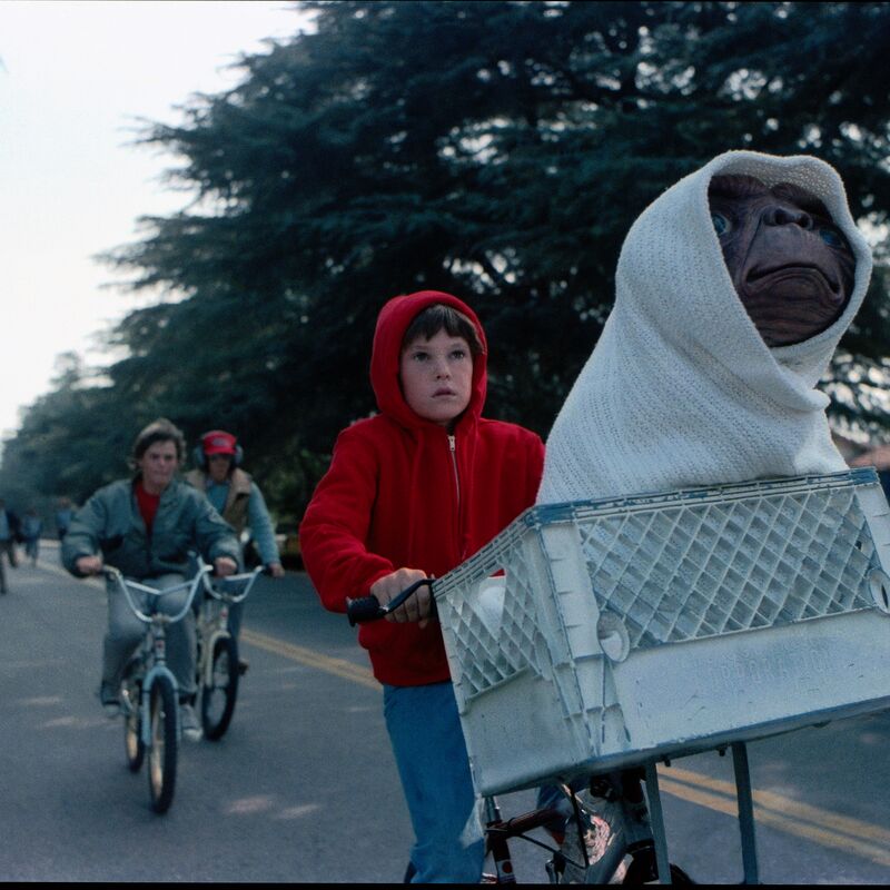 A frame from the film ET featuring Elliot wearing a red hoodie cycling ET, who's covered with a white towel and sits in Elliot's bike's basket, around